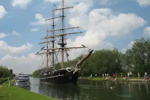 Historisches Segelschiff am Fluss mit grüner Uferwiese, daneben ein Boot und Menschen am Steg.