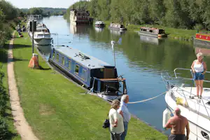 Blaue Hausbootanlage am Kanal: Liegeplätze mit mehreren Booten, grüner Uferpromenade und Bäumen entlang des Wassers.