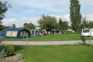 Campingplatz mit Wohnwagen und Zelten, grüner Rasenfläche, Kiesweg und Bäumen im Hintergrund.