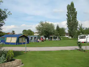 Campingwiese mit mehreren Zelten und Wohnwagen, daneben ein Auto und ein Weg zwischen den Stellplätzen, umgeben von Bäumen.