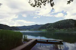 Holzsteg am See mit Blick auf ruhiges Wasser, Schilf am Ufer und bewaldete Hügel in der Ferne.