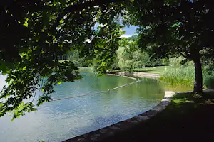 Ruhiger See am Campingplatz, Uferweg und Stege, Bäume spenden Schatten und Schilf wächst am Rand.