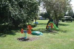Spielplatz auf dem Campingplatz: Federwippe und Rutsche im Gras, Bäume und Wohnwagen im Hintergrund.