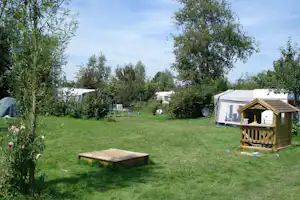 Campingwiese mit mehreren Zelten, Gartenmöbeln und einem kleinen Holz-Spielhaus im Vordergrund, umgeben von Bäumen.