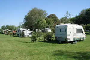 Wohnwagen auf einem grünen Campingplatz, daneben weitere Fahrzeuge und Zelte hinter Büschen und Bäumen.