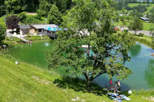 Campingplatz am See mit Holzstegen, Terrasse und Bootssteg, umgeben von Bäumen und Wiesen.