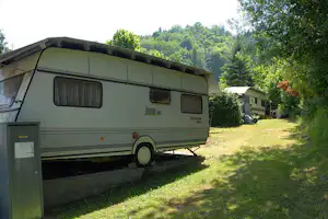 Wohnwagen auf einem Campingplatz-Stellplatz, mit Grasfläche, Hecken und Bäumen im Hintergrund.