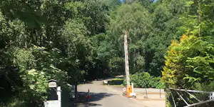 Einfahrt zum Campingplatz mit Asphaltweg, Schranke und Bäumen im Grünen, daneben ein Schild und Absperrpfosten.