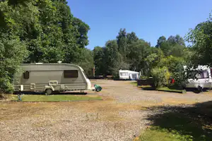 Wohnwagen auf einem Campingplatz-Stellplatz, daneben ein weiterer Caravan und ein Wohnmobil, umgeben von Bäumen und Grün.