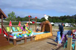 Aufblasbares Spielbecken mit Holzpodest, bunten Luftballons und einem Vogel-Dekor auf dem Campingplatz, im Hintergrund Bungalows und Bäume.