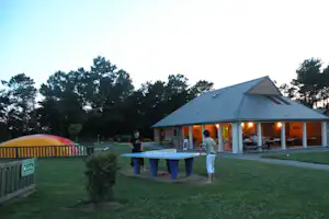 Campinghaus mit beleuchteter Terrasse, davor ein Tisch und Stühle, im Hintergrund Bäume und ein Spielbereich mit buntem Zelt.