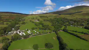 Luftaufnahme eines Campingplatzes im grünen Tal mit vielen Stellplätzen, Wegen und umliegenden Hügeln unter blauem Himmel.
