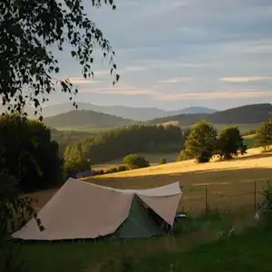 Zelt auf einer Wiese im Grünen, im Hintergrund Hügel und Felder, Bäume im Vordergrund bei Abendlicht.