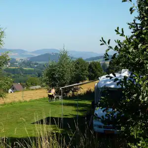 Wohnwagen auf einer Campingwiese mit Blick ins Tal und auf bewaldete Berge, umgeben von Büschen und Gras.