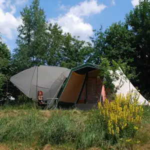 Zelt auf einem Campingplatz im Grünen, mit offenem Vorzelt und Sitzgelegenheit am Rand zwischen Büschen und Bäumen.