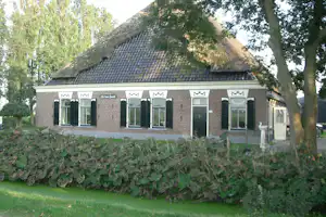 Ziegelhaus mit Reetdach und großen Fenstern, davor Rasen und Hecke, im Hintergrund Bäume.