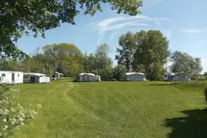 Grüner Campingplatz mit Rasenweg, mehreren Wohnwagen und Mobilheimen, umgeben von Bäumen und einem weißen Gebäude im Hintergrund.