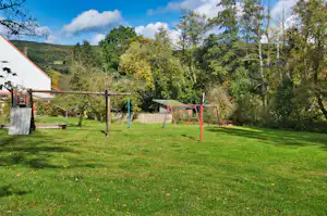 Grüne Campingwiese mit Spielgeräten, Schaukel und Rutsche vor einem weißen Gebäude und Bäumen am Waldrand.