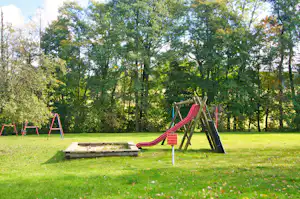 Spielplatz am Campingplatz: Holzpodest und Rutsche mit Schaukel, umgeben von Rasen und Bäumen im Hintergrund.