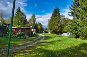 Campingweg mit grünem Rasen, Bäumen und Hecken, links ein Gebäude und rechts Stellplätze mit Wohnwagen.
