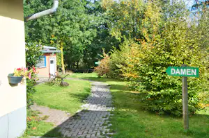 Gepflasterter Campingweg durch den Garten mit Büschen und Schild „DAMEN“ neben einem Gebäude.