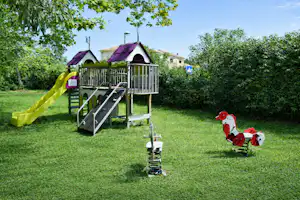 Spielplatz im Grünen mit Klettergerüst und Rutsche, daneben ein kleines Spielhaus und ein rotes Spielzeugpferd.