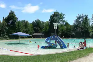 Blaues Planschbecken mit Rutsche, Sonnenschirm und Liegefläche am Campingplatz, im Hintergrund Bäume und Gebäude.