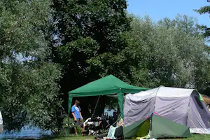 Grüne Campingzelt mit Vorzelt und weißem Zelt daneben, Campingausrüstung am Boden, umgeben von Bäumen im Schatten.
