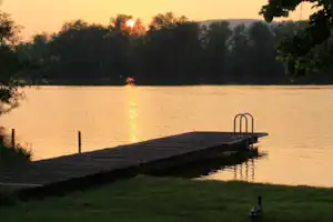 Holzsteg am See am Sonnenuntergang, mit Leiter und Geländer, im Hintergrund Bäume und Uferlinie.
