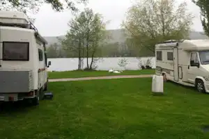 Wohnmobile auf einem grünen Campingplatz am See, Bäume im Hintergrund und ein Tisch mit Wäschekorb am Stellplatz.
