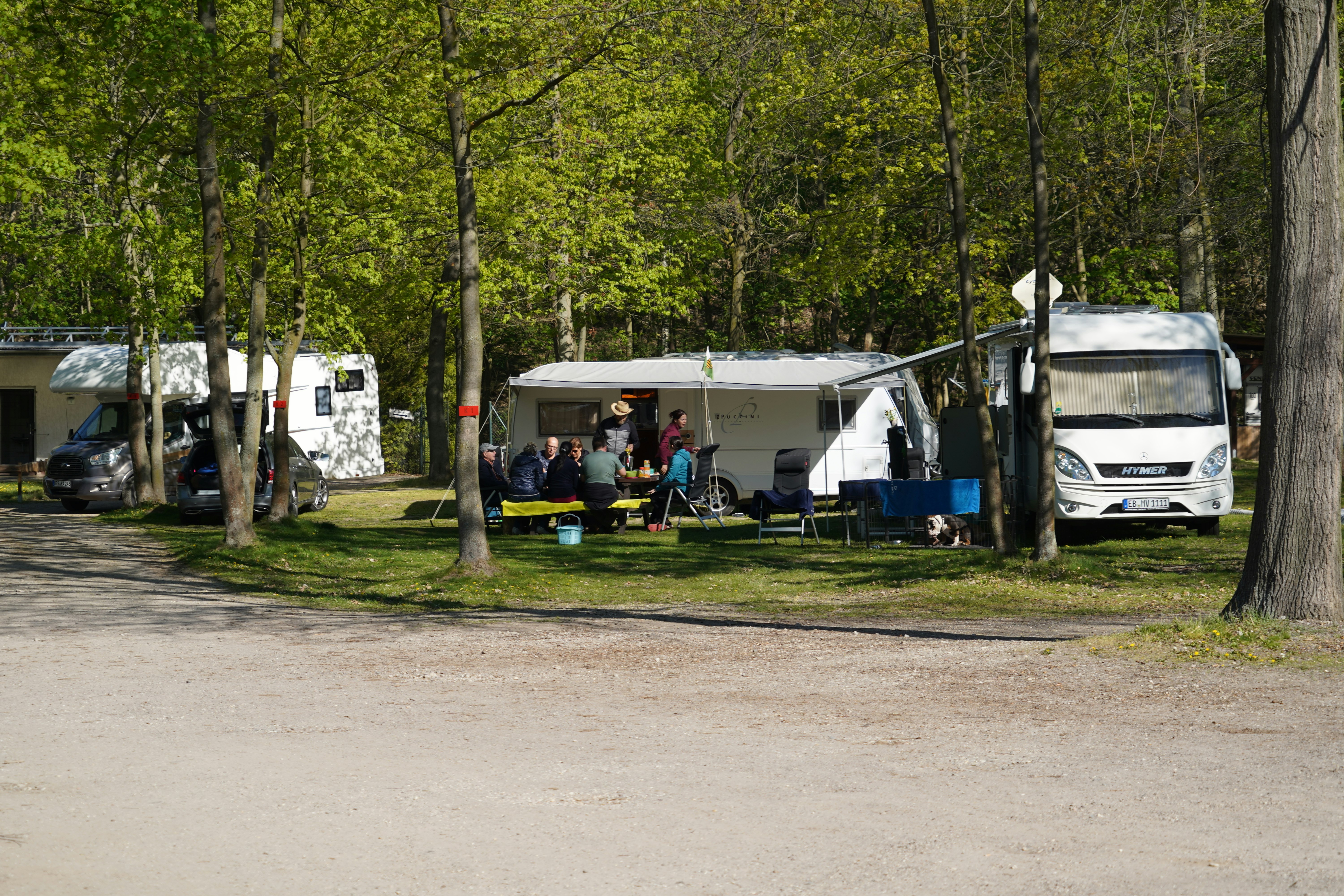 Wohnwagen und Campingbus auf einem Stellplatz zwischen Bäumen, mit Tisch und Stühlen im Vordergrund.