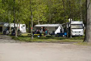Wohnwagen und Campingbus auf einem Stellplatz zwischen Bäumen, mit Tisch und Stühlen im Vordergrund.
