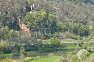 Blick ins Tal mit dichtem Wald, Fluss und Wiesen, im Hintergrund Hügel und ein kleines Gebäude am Hang.