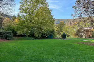 Grüner Rasen mit Bäumen und Blick auf ein Gebäude in der Ferne, umgeben von Hecken und Wegen.