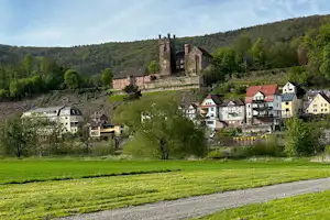 Grünes Tal mit Wiese und Kiesweg, im Hintergrund ein Dorf und eine Burg auf dem Hügel.