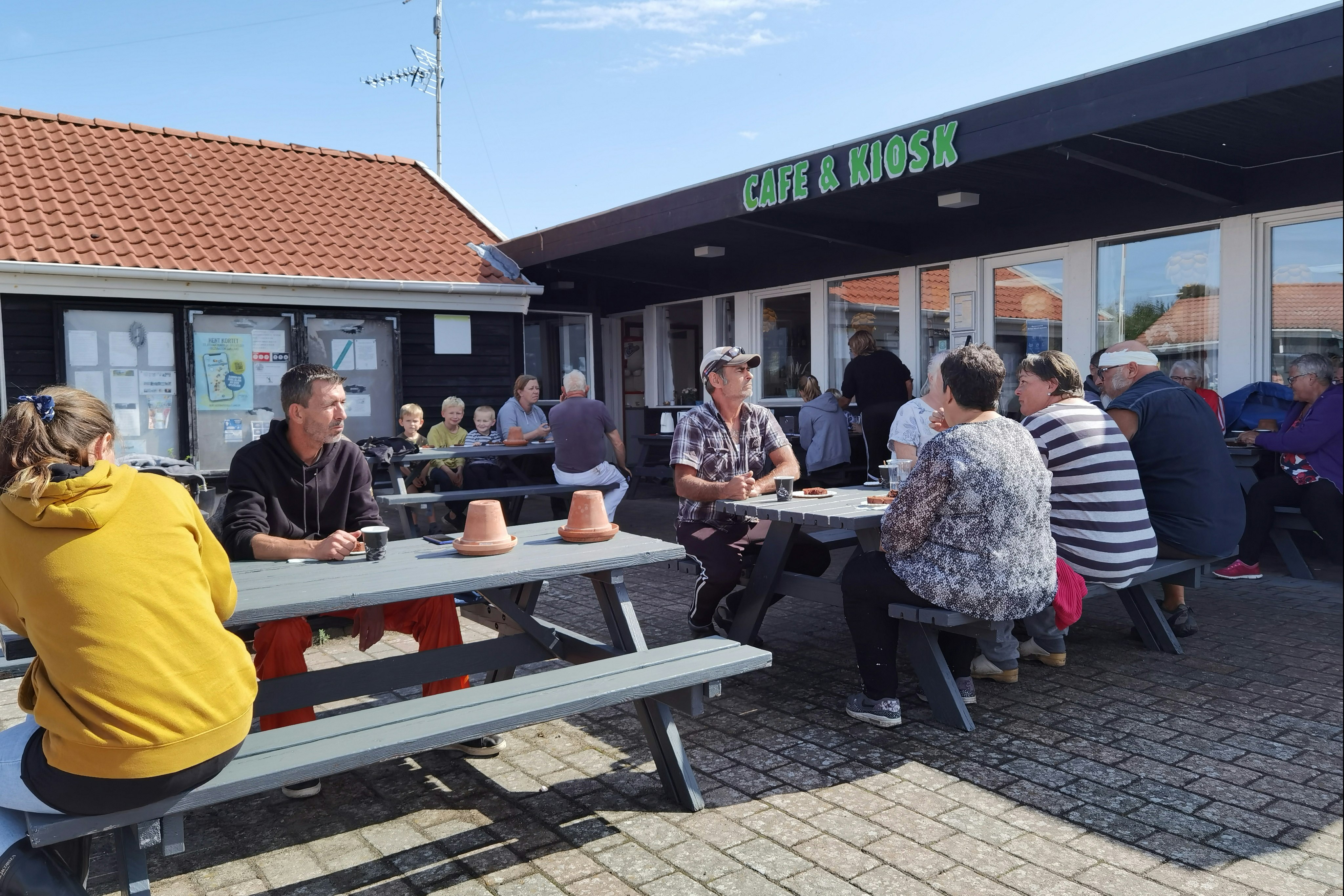 Café & Kiosk am Campingplatz mit Sitzgruppen an Holztischen, Menschen im Außenbereich vor dem Gebäude.