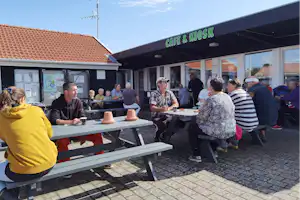 Café & Kiosk am Campingplatz mit Sitzgruppen an Holztischen, Menschen im Außenbereich vor dem Gebäude.