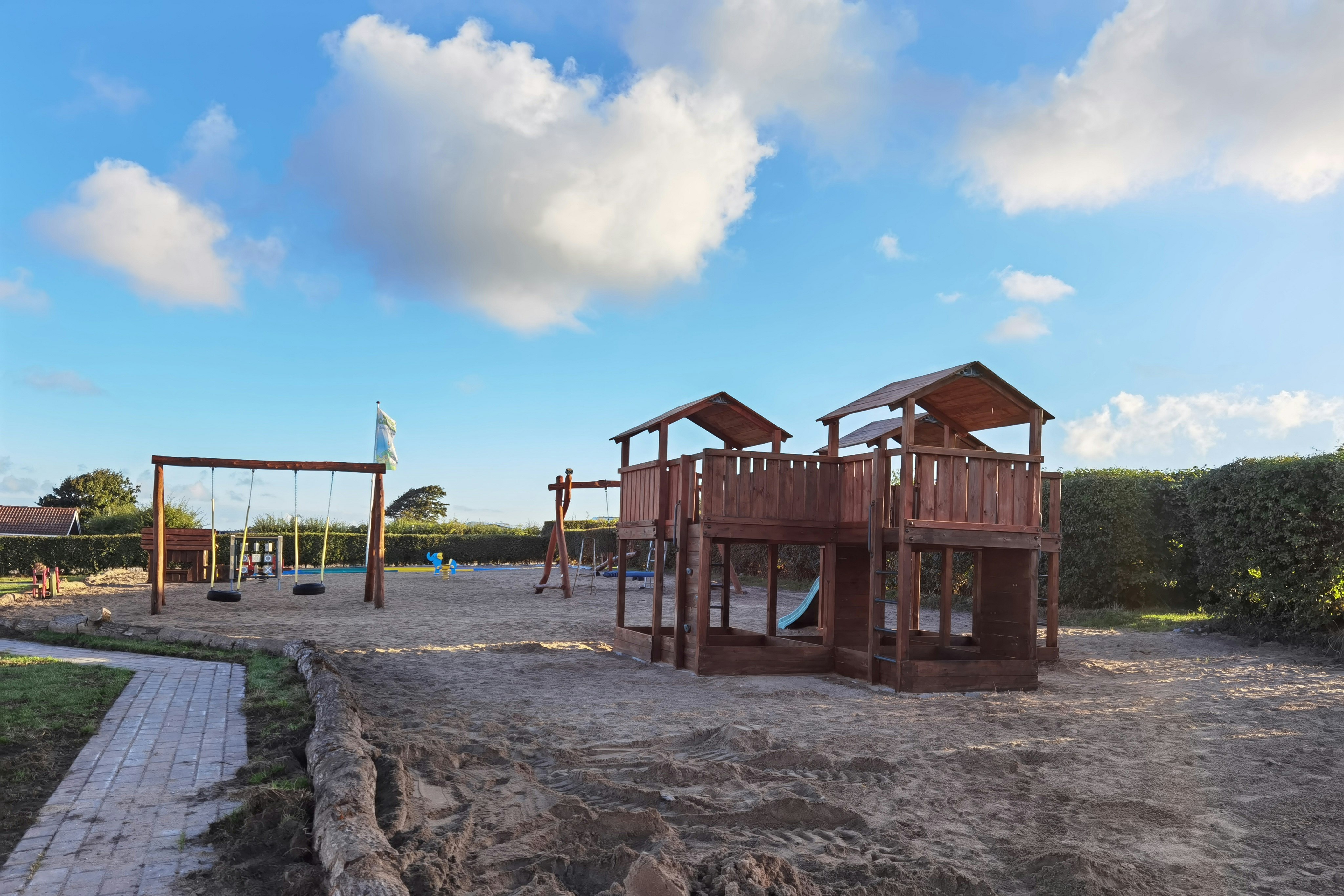 Spielplatz am Campingplatz mit Holzspielgeräten, Sandfläche und Zaun, im Hintergrund Wasser und Himmel.