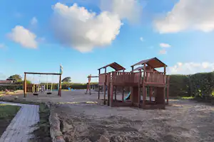 Spielplatz am Campingplatz mit Holzspielgeräten, Sandfläche und Zaun, im Hintergrund Wasser und Himmel.