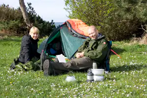 Zelt im Grünen auf einem Campingplatz, zwei Personen sitzen davor auf einer Decke, daneben Campinggeschirr und Getränke.