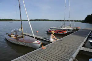 Segelboote am Steg, eine Person sitzt auf dem Holzsteg am See, im Hintergrund Wald und Wasser.