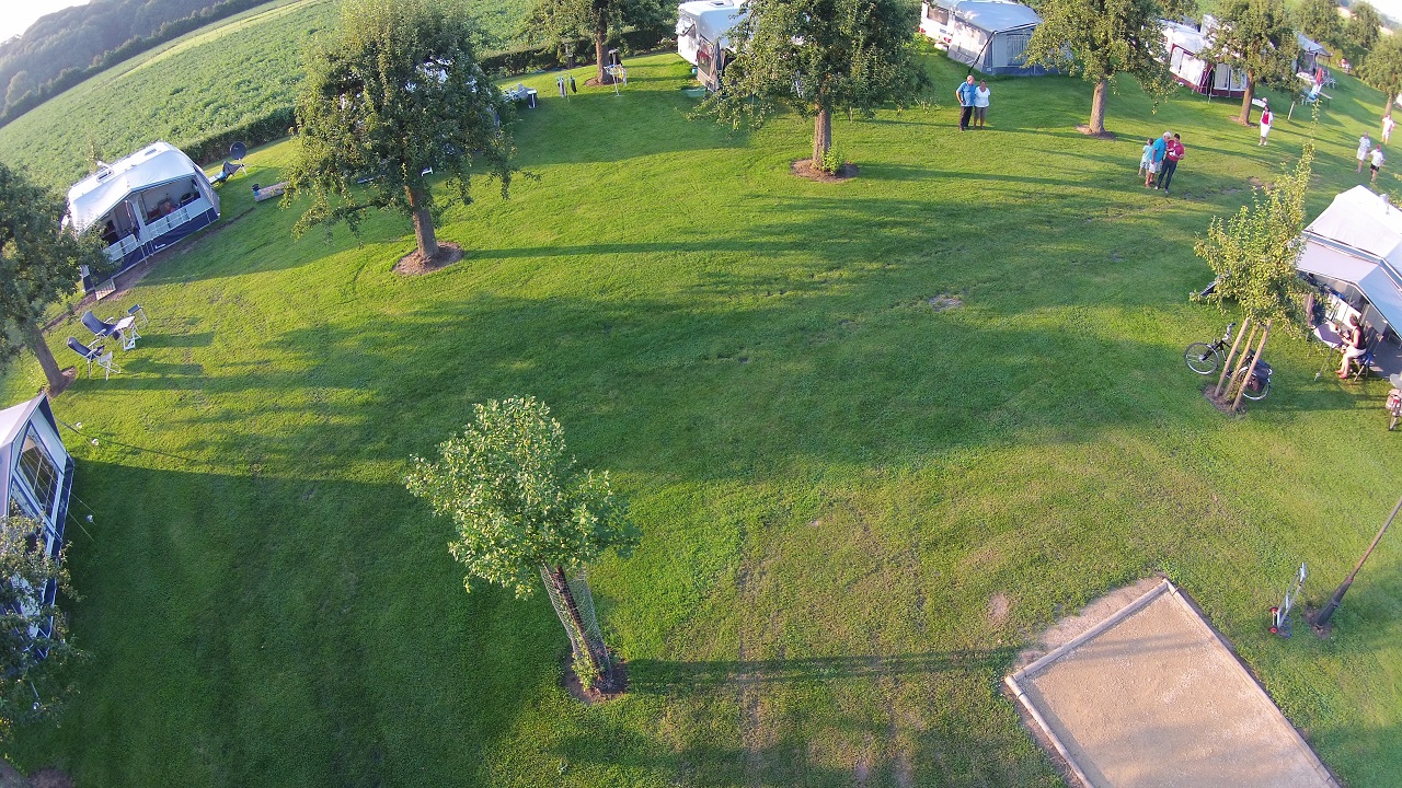 Campingplatz mit Rasenfläche, Wohnwagen und Zelten, Bäumen und Wegen; im Hintergrund Häuser und ein Spielbereich.