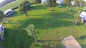 Campingplatz mit Rasenfläche, Wohnwagen und Zelten, Bäumen und Wegen; im Hintergrund Häuser und ein Spielbereich.