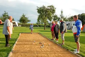 Boule-Spiel auf einem Campingplatz: mehrere Personen stehen am Bahnrand, im Hintergrund Zelte und Bäume auf der Wiese.
