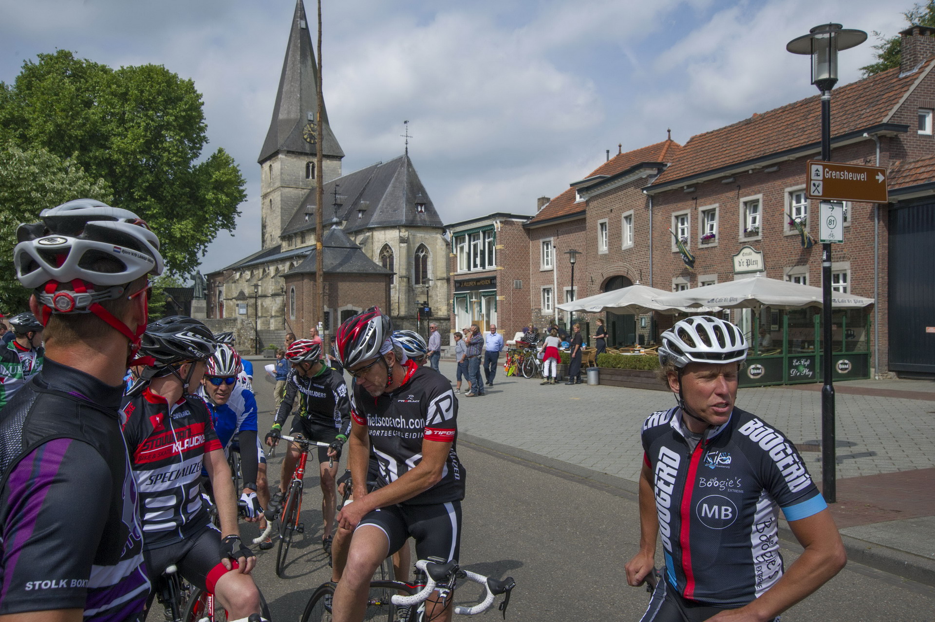 Radfahrer auf einem Platz vor einer Kirche, mit Fahrrädern und Helmen, daneben Häuser und ein Straßenschild.