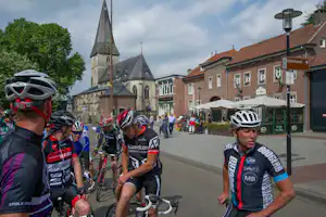 Radfahrer auf einem Platz vor einer Kirche, mit Fahrrädern und Helmen, daneben Häuser und ein Straßenschild.