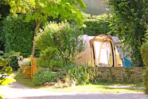 Zelt auf einem Campingplatz auf einem gepflasterten Stellplatz, umgeben von Büschen, Blumen und einem Steinwall.