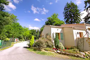 Ferienhaus mit Terrasse und Eingang am Campingplatz, umgeben von Hecken, Blumenbeeten und Bäumen entlang eines Kieswegs.