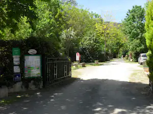 Eingang eines Campingplatzes mit Weg, Infotafeln und Schranke, umgeben von Bäumen und grünem Gelände.
