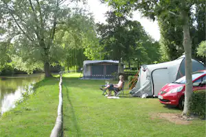 Campingplatz am Fluss: Zelte und ein rotes Auto am Stellplatz, daneben ein Tisch und Stühle im Grünen.